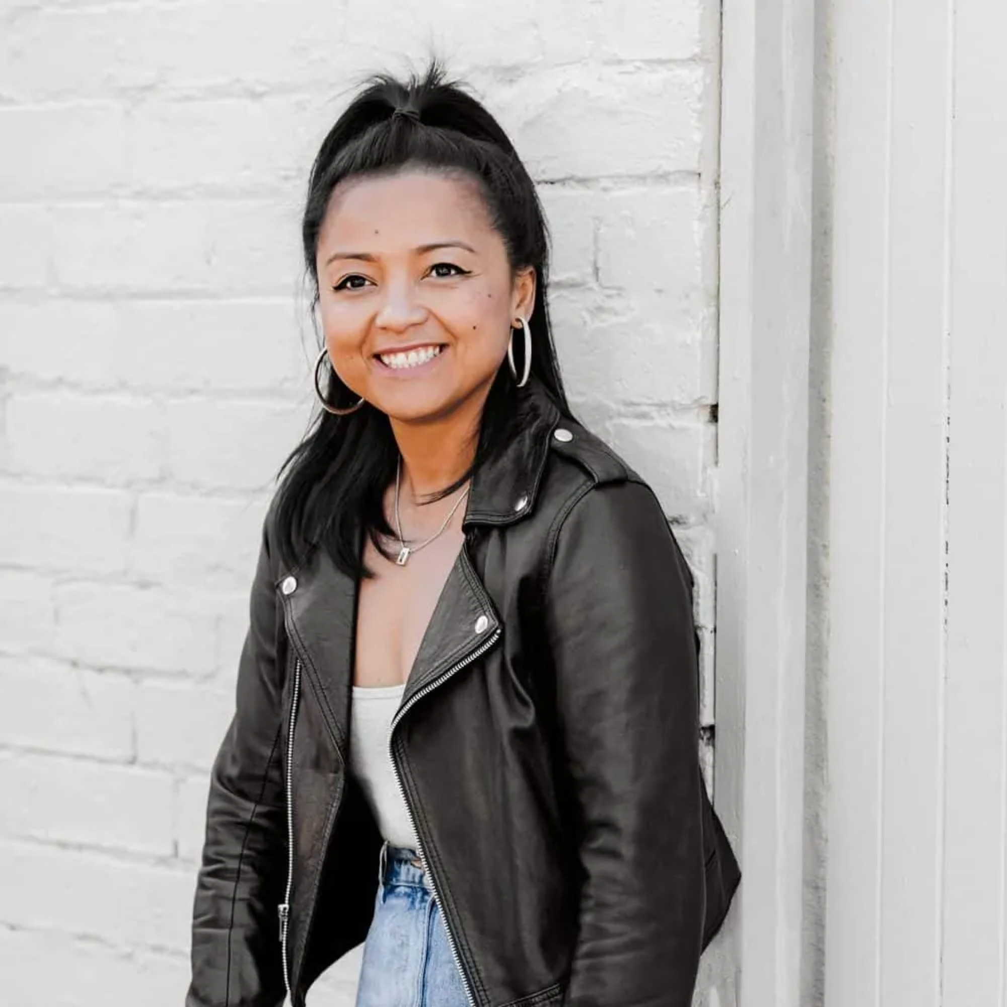 smiling person wearing a black leather jacket and blue jeans leans against a white wall