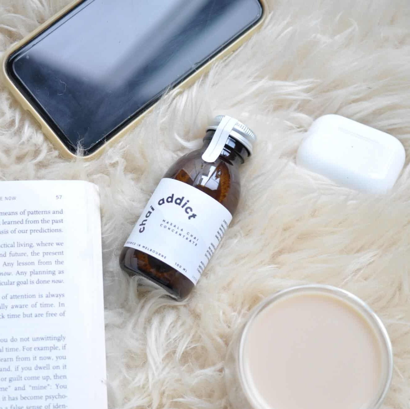 100ml chai addict logo on top of a warm and comforting shaggy rug with a book, cup of hot chai, airpods and iphone