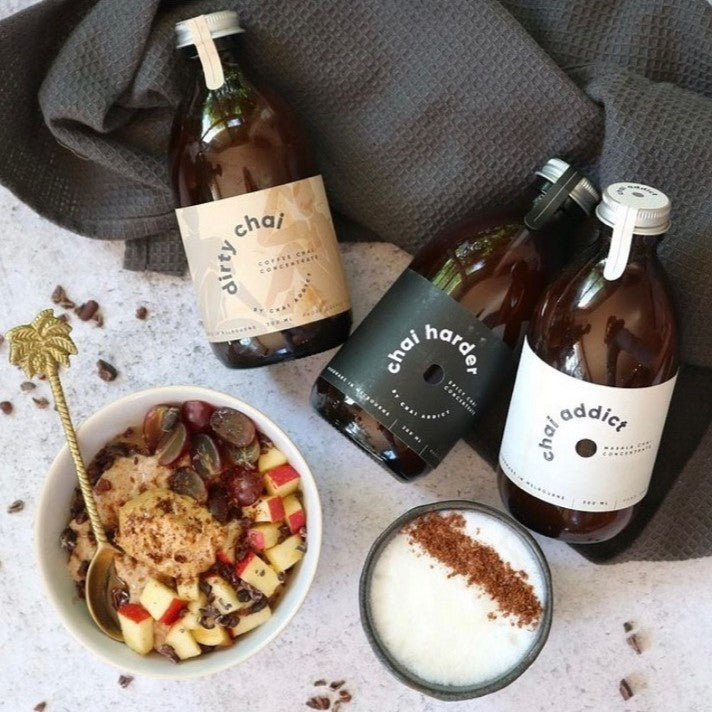 a bowl of apple crumble with a gold teaspoon is served alongside a cup of hot chai latte, along with 3 bottles of chai latte concentrates by chai addict