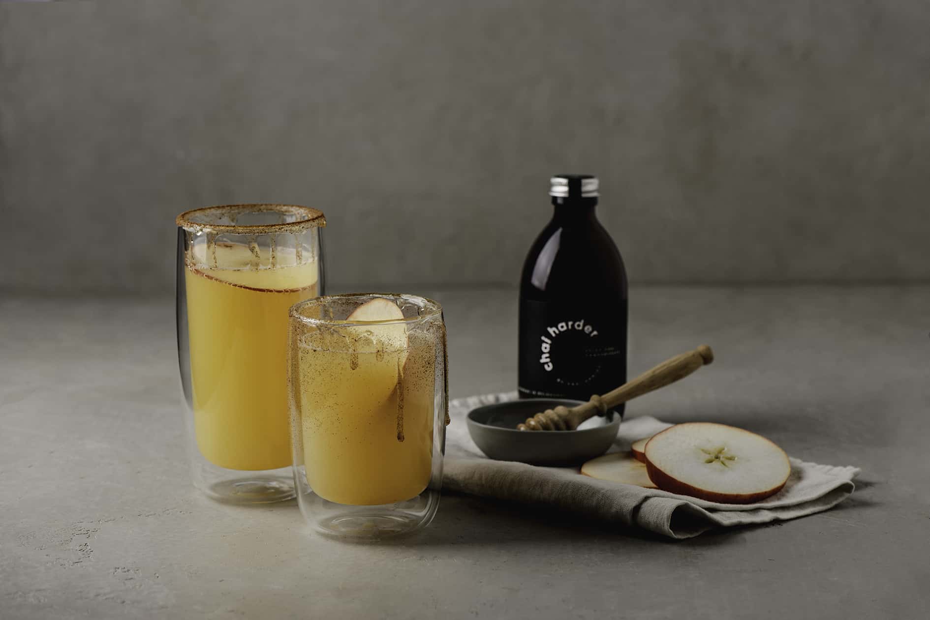 refreshing chai soda served in a glass tumbler dripping with honey, a small saucer with honey wand and round sliced apples on grey tea towel and a bottle of chai harder spicy chai concentrate