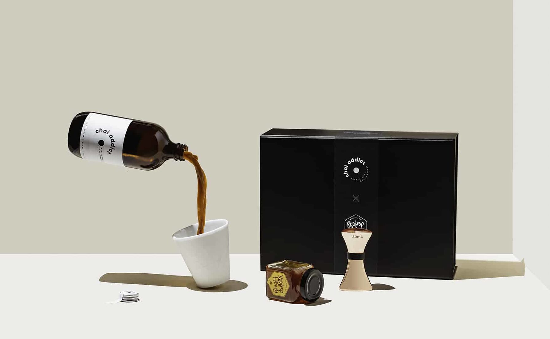 levitating chai addict masala chai concentrate pouring in a white ceramic cup next to a black box, jar of honey and jigger